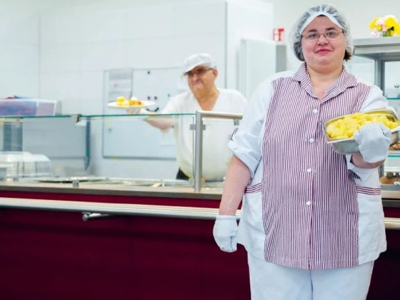 Frau in wei&szlig;er Arbeitskleidung und gestreifter rot-wei&szlig;er Sch&uuml;rze h&auml;lt eine Metallschale mit Kartoffeln und tr&auml;gt Handschuhe sowie eine Haube. Im Hintergrund ein Mann in &auml;hnlicher Kleidung mit einem Teller; dahinter eine Essensausgabe mit Glasabdeckung. Wand und Einrichtung in Wei&szlig; gehalten.
