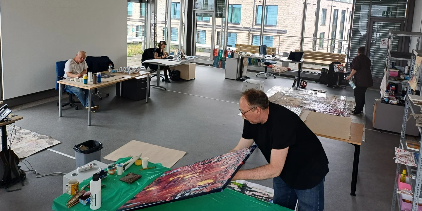 Gro&szlig;er, lichtdurchfluteter Raum mit Panoramafenstern. Vier Personen besch&auml;ftigt an unterschiedlichen Tischen: Im Vordergrund ein Mann, der ein buntes Gem&auml;lde auf einem gr&uuml;nen Tisch bearbeitet. Links zwei Personen an separaten Tischen, rechts eine weitere Person an einem gro&szlig;en Arbeitsplatz. Auf den Tischen Malutensilien und Werkzeuge. Hintergrund zeigt moderne B&uuml;rogeb&auml;ude.