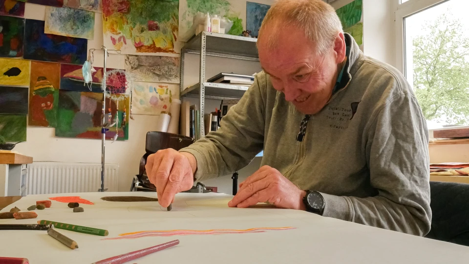 Ein &auml;lterer Mann in grauem Pullover zeichnet mit einem orangefarbenen Stift auf wei&szlig;em Papier. Auf dem Tisch liegen bunte Stifte und Pastelle verstreut. Im Hintergrund ein Regal mit B&uuml;chern und Materialien sowie eine bunte Wand mit abstrakten Kunstwerken. Tageslicht f&auml;llt durch ein gro&szlig;es Fenster rechts ein.
