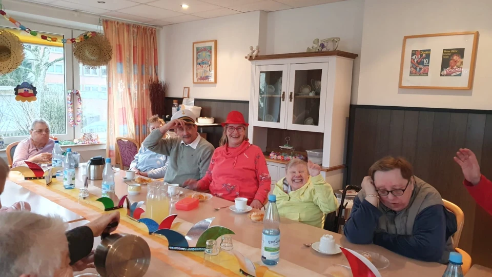 Langer Tisch mit buntem, dekorativem Streifen und Origami-Elementen, gedeckt mit wei&szlig;en Tassen, Gl&auml;sern, Wasserkaraffen, Saftkrug, Kaffeekanne und Snacks. Im Hintergrund ein Holzschrank mit Geschirr, helle Gardinen, Fenster mit Blick auf B&auml;ume, bunte Wandbilder.