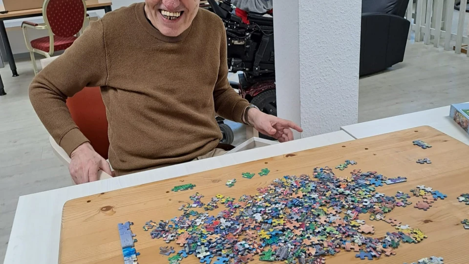 Ein Holzbrett mit einem Haufen bunter Puzzleteile auf einem wei&szlig;en Tisch. Im Hintergrund ein heller Raum mit roten und schwarzen St&uuml;hlen, einem Rollstuhl und hohen Fenstern mit Blick ins Gr&uuml;ne.