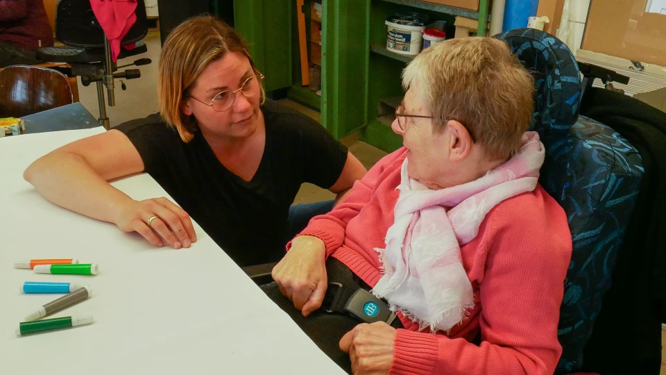 Zwei Personen an einem wei&szlig;en Tisch in einem Arbeits- oder Gemeinschaftsraum, links eine Person in schwarzem T-Shirt, rechts eine Person im Rollstuhl mit rosa Pullover und wei&szlig;em Schal. Auf dem Tisch liegen farbige Filzstifte, im Hintergrund sind Regale, Arbeitsmaterialien und St&uuml;hle sichtbar.