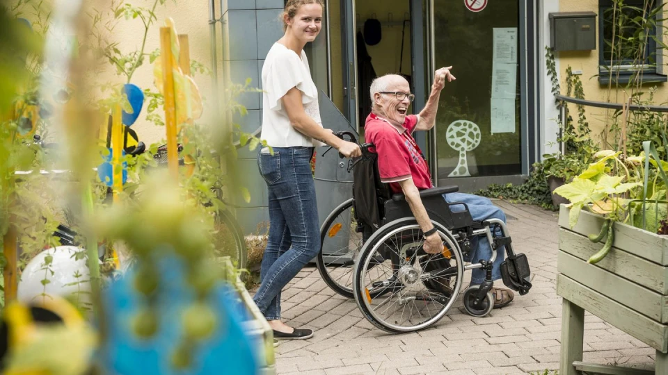 Eine Pflegerin schiebt einen Senioren im Rollstuhl und beide l&auml;cheln in die Kamera.