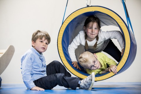 Kinder spielend in einer Sporthalle - zwei liegen in einem Spieltunnel, ein Kind sitzt davor