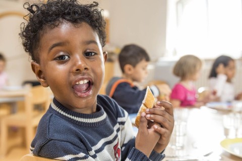 Ein Junge mit kurzen Locken isst fröhlich sein Baguette, im Hintergrund sind weitere Kinder zu sehen