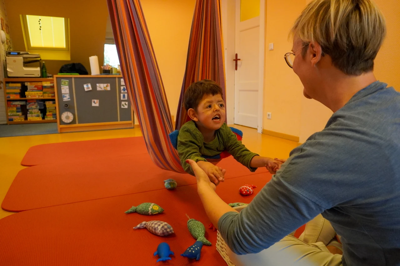 Ein Kind sitzt auf einem orangefarbenen Gymnastikmattenboden in einem Raum mit gelb gestrichenen W&auml;nden. Es liegt in einer rot gestreiften H&auml;ngeschaukel und h&auml;lt die H&auml;nde eines Erwachsenen mit kurzen, blonden Haaren. Auf dem Boden liegen mehrere bunte, stoffartige Spielzeuge in Fisch- und Tierformen verteilt. Im Hintergrund eine graue Kommode mit Aufklebern, Regale mit Spielsachen und eine wei&szlig;e T&uuml;r.