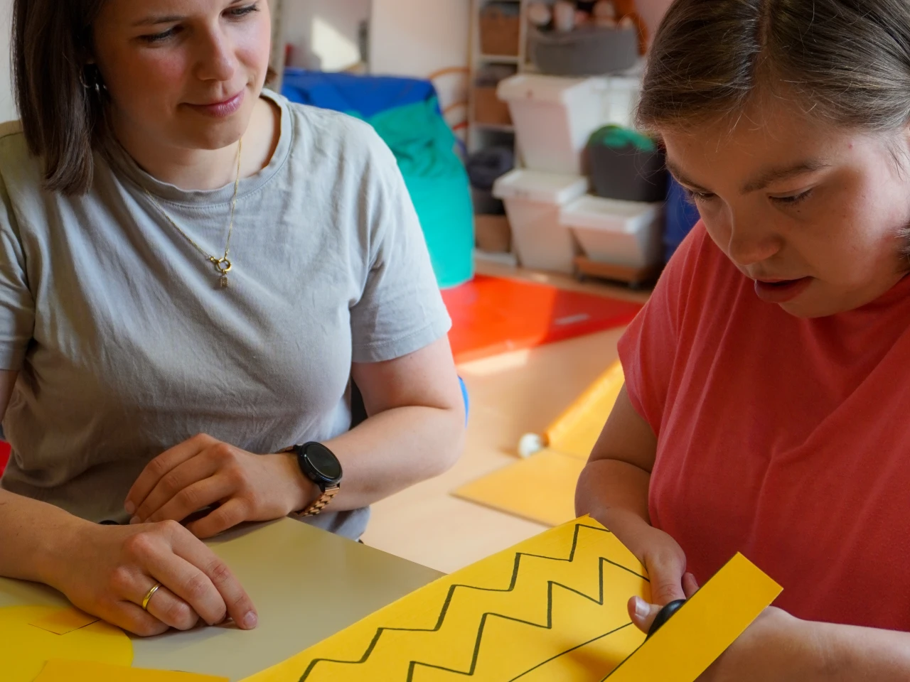 Zwei Personen sitzen an einem Tisch in einem Raum mit leuchtend orangefarbenen und gelben Gymnastikb&auml;llen im Hintergrund. Auf dem Tisch liegt gelbes Papier mit schwarzen Zickzacklinien; eine Person h&auml;lt eine Schere, um das Papier entlang der Linien zu schneiden.