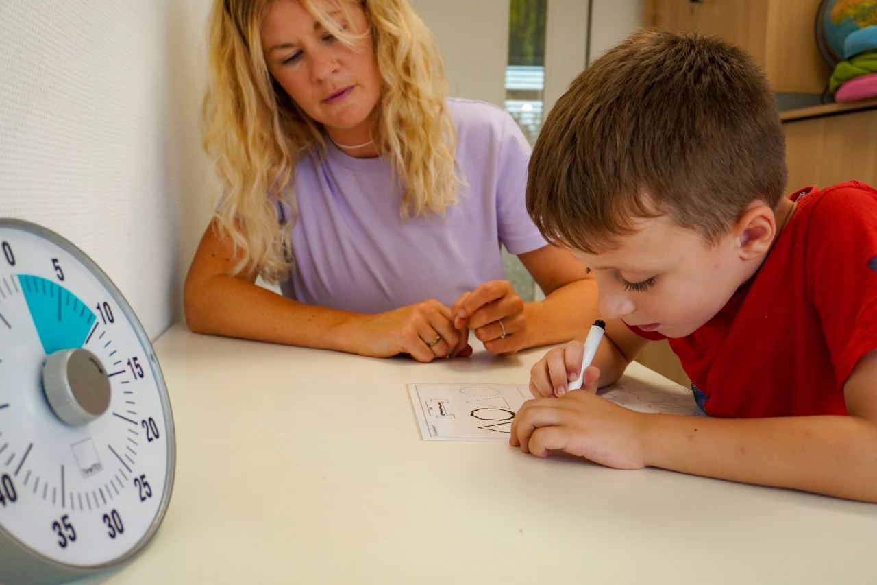 Ein Kind in rotem T-Shirt schreibt mit einem wei&szlig;en Stift auf einem Arbeitsblatt auf einem Tisch. Neben ihm liegt eine Eiersanduhr mit Minutenmarkierungen. Im Hintergrund eine helle Wand und ein Schrank mit buntem Spielzeug.