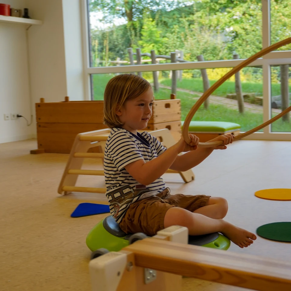 Kind in gestreiftem T-Shirt und brauner Shorts sitzt auf einer gr&uuml;nen, halbkugelf&ouml;rmigen Wippe und h&auml;lt zwei Holzreifen. Raum mit hellem Boden, Holzspielm&ouml;beln, bunten Matten und gro&szlig;em Fenster mit Blick auf gr&uuml;ne Wiese und B&auml;ume.