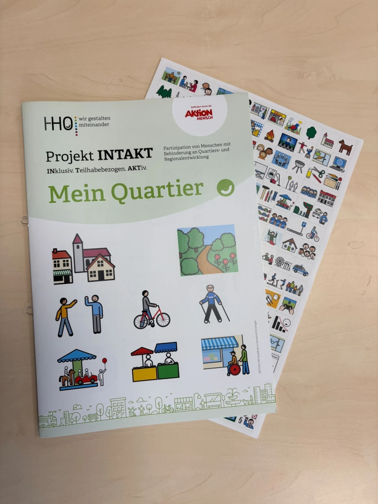 Heft mit dem Titel "Projekt INTAKT &ndash; INklusiv. Teilhabebezogen. AKTiv. Mein Quartier" auf hellem Holztisch, daneben ein Blatt mit Symbolen und Illustrationen. Das Heft enth&auml;lt Zeichnungen von H&auml;usern, B&auml;umen, Personen in Alltagssituationen, St&auml;nden und Gesch&auml;ften. Oben rechts das Logo "Aktion Mensch".