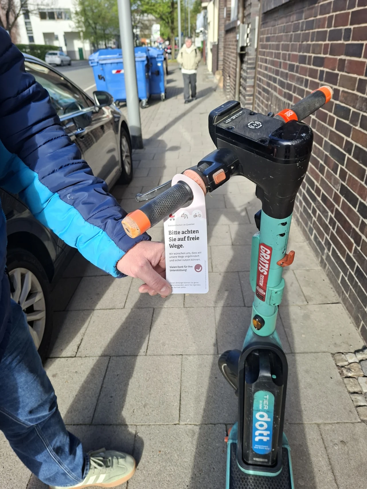 Elektroroller in t&uuml;rkis und schwarz mit orangefarbenen Griffen, geparkt auf einem B&uuml;rgersteig. Am Lenker h&auml;ngt ein Schild mit der Aufschrift: "Bitte achten Sie auf freie Wege. Wir m&ouml;chten sicher sein, dass sich alle sicher f&uuml;hlen. Vielen Dank f&uuml;r Ihre R&uuml;cksichtnahme." Links h&auml;lt eine Person in blauer Jacke das Schild. Im Hintergrund Gehweg, parkendes Auto, blaue M&uuml;lltonnen und weitere Personen.