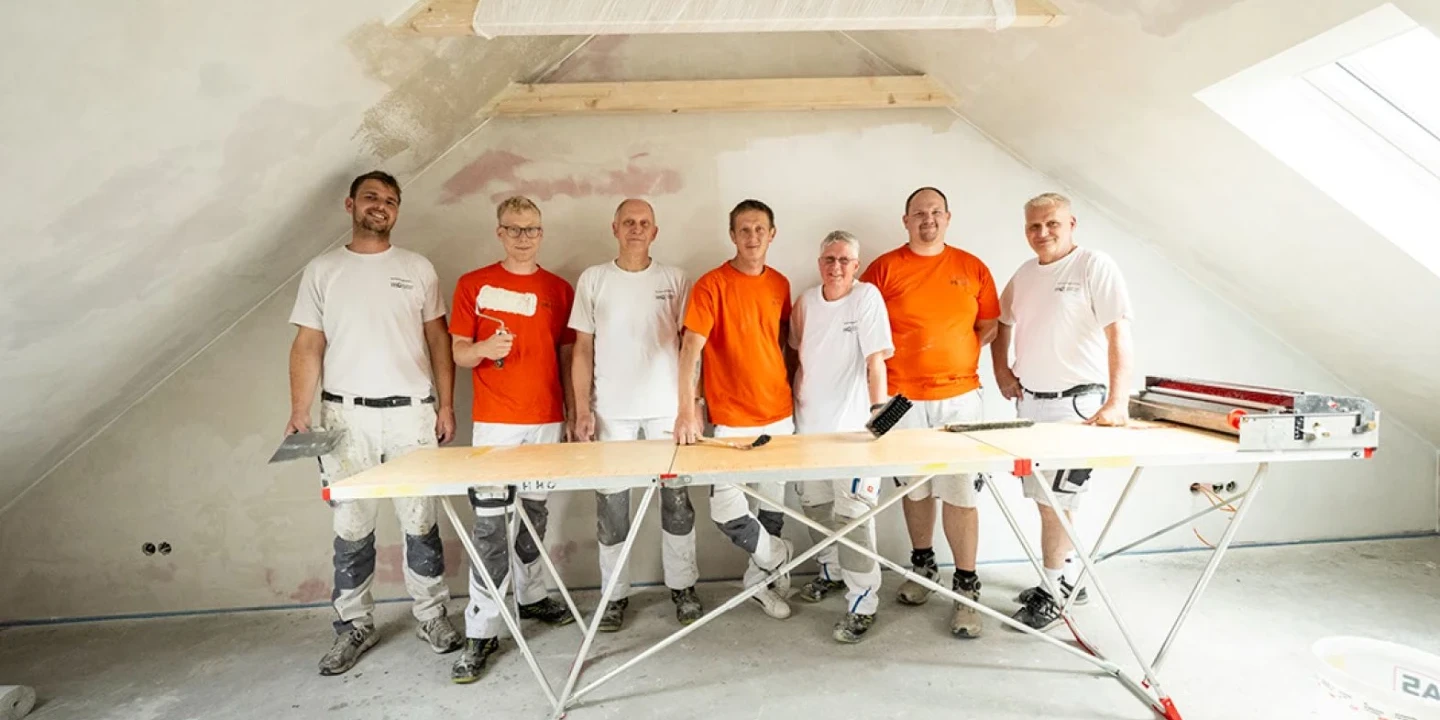 Sechs Personen in Arbeitskleidung stehen nebeneinander in einem fast leer renovierten Raum mit schr&auml;gen W&auml;nden. Zwei tragen wei&szlig;e, vier orange Oberteile. Vor ihnen ein Arbeitstisch mit Werkzeugen, darunter ein Ma&szlig;band und ein Kunststoffhalter. Links und rechts im Bild unfertige W&auml;nde, rechts oben ein Fenster.