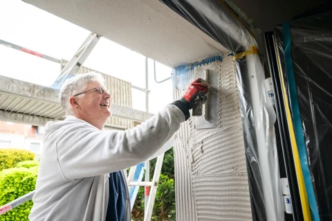 &Auml;lterer Mann mit grauen Haaren und Brille tr&auml;gt in einem wei&szlig;en Pullover und roten Arbeitshandschuhen Putz mit einer Gl&auml;ttkelle auf eine mit Gewebe bedeckte Wand auf. Im Hintergrund sind eine Leiter, ein Bauger&uuml;st und gr&uuml;ne B&uuml;sche sichtbar.