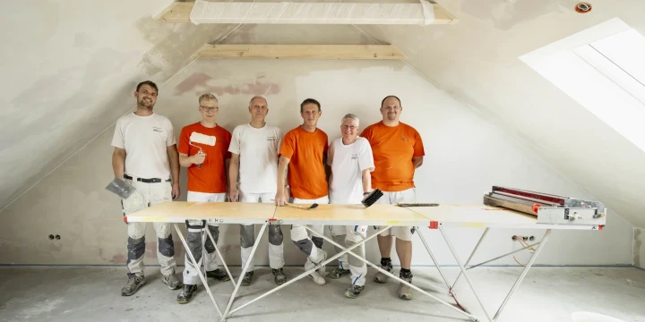 Sechs Personen in Arbeitskleidung stehen nebeneinander in einem fast fertig renovierten Dachgeschossraum mit schr&auml;gen W&auml;nden und einer wei&szlig;en Decke. Vier Personen tragen wei&szlig;e Oberteile, zwei tragen orangefarbene Oberteile. Vor ihnen ein Baustellentisch auf einem Ger&uuml;st, darauf Werkzeuge und eine Malerrolle. Rechts ein Schneideger&auml;t f&uuml;r Gipsplatten. W&auml;nde teils verspachtelt.