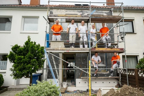 Reihenhaus mit wei&szlig;er Fassade und rotem Ziegeldach, an dem ein Bauger&uuml;st angebracht ist. Auf dem Ger&uuml;st stehen sechs Arbeiter in orangenen und wei&szlig;en Arbeitskleidern, teils l&auml;chelnd. Links ein Baum, rechts ein Zaun; im Vordergrund Erde und Gehwegplatten.