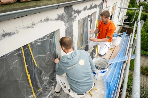Zwei M&auml;nner arbeiten auf einem Ger&uuml;st an der Fassade eines Geb&auml;udes. Die Fenster sind mit transparenter Folie und gelbem Klebeband abgedeckt. Der Mann links tr&auml;gt eine graue Kapuzenjacke, der rechts ein oranges T-Shirt. Neben ihnen stehen wei&szlig;e Farbeimer und Werkzeuge.