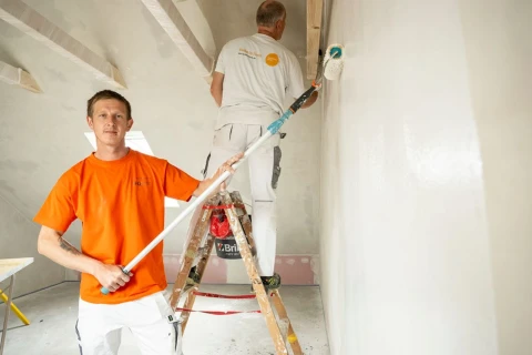 Zwei M&auml;nner streichen eine helle Wand in einem Raum mit Dachschr&auml;gen. Der vordere Mann in orangefarbenem T-Shirt und wei&szlig;er Arbeitskleidung h&auml;lt eine Farbrolle mit Verl&auml;ngerungsstange. Der hintere Mann steht auf einer Holzleiter und streicht die Wand mit einer kurzen Farbrolle. Die Decke hat sichtbare wei&szlig;e Balken, und links ist ein Fenster sichtbar.
