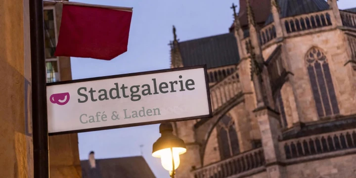 Schild mit der Aufschrift &bdquo;Stadtgalerie Caf&eacute; & Laden&ldquo; an einem Laternenpfahl mit warm leuchtender Stra&szlig;enlaterne. Daneben eine rote Fahne. Im Hintergrund eine gro&szlig;e, gotische Kirche mit Spitzbogenfenstern und filigranen T&uuml;rmen.