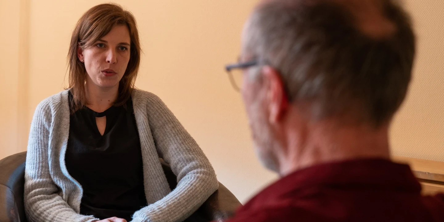 Eine Frau mit schulterlangem braunem Haar, schwarzem Oberteil und grauer Strickjacke sitzt in einem schwarzen Stuhl und blickt ernsthaft nach vorne. Im Vordergrund sitzt ein &auml;lterer Mann mit kahler Stelle und Brille, von hinten sichtbar, in einem roten Hemd. Hintergrund in beige.