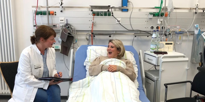 Eine &Auml;rztin spricht mit einer lachenden Frau, die im Krankenbett liegt.