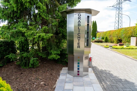 Silberner Automat mit der Aufschrift "Grablichter", aufgestellt auf einem mit grauen Pflastersteinen gestalteten Weg. Im Automaten sind zul&auml;ssige Grablichter sichtbar. Links davon ein Beet mit Erde und einer gro&szlig;en Tanne, rechts Hecken, Rasenfl&auml;chen und Stromleitungsmasten.
