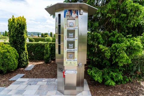 Automat aus Edelstahl mit f&uuml;nf Wasserbeh&auml;ltern unterschiedlicher Gr&ouml;&szlig;e und Preisen von 2,00 bis 3,50 Euro. Oben zwei Fotos von Personen, darunter die Beschriftung "H₂O". Auf der linken Seite gr&uuml;ne Hecken und B&auml;ume, rechts eine gro&szlig;e Tanne. Der Automat steht auf einem gepflasterten Untergrund.
