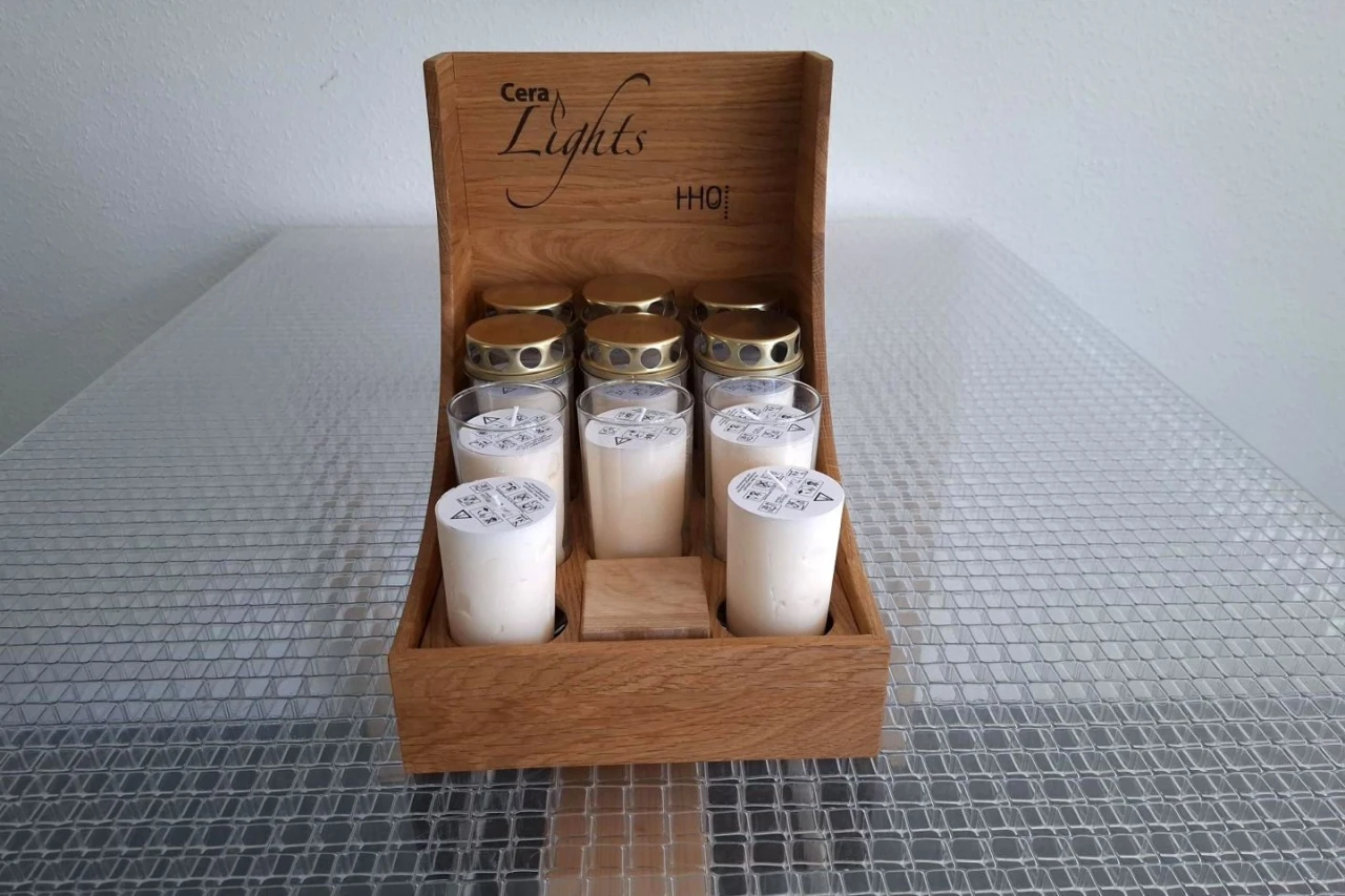 Holzgestell mit der Aufschrift "Cera Lights" und "HQ!" enth&auml;lt sechs wei&szlig;e Kerzen in Glasbeh&auml;ltern mit goldfarbenen Deckeln und vier wei&szlig;e Stumpenkerzen mit bedruckten Papierabdeckungen. Platte Oberfl&auml;che mit gitterartigem Muster darunter.