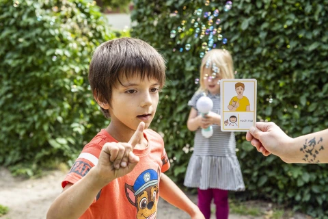 Kind in rotem T-Shirt mit Paw-Patrol-Motiv zeigt mit dem Finger, daneben eine Hand mit bebilderter Karte "noch mal". Im Hintergrund ein weiteres Kind im gestreiften Kleid h&auml;lt einen Seifenblasenstab. Gr&uuml;nen Hecken und schwebende Seifenblasen bilden den Hintergrund.