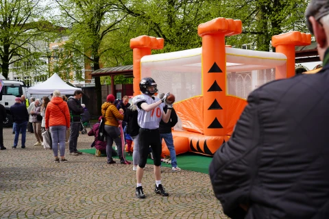 Person in American-Football-Ausr&uuml;stung mit schwarzem Helm und Trikot mit der Nummer 70 h&auml;lt einen Football in Wurfposition, daneben orangefarbene H&uuml;pfburg mit schwarzen Dreiecken, umgeben von sitzenden und stehenden Menschen auf einem gepflasterten Platz mit gr&uuml;nen B&auml;umen im Hintergrund.