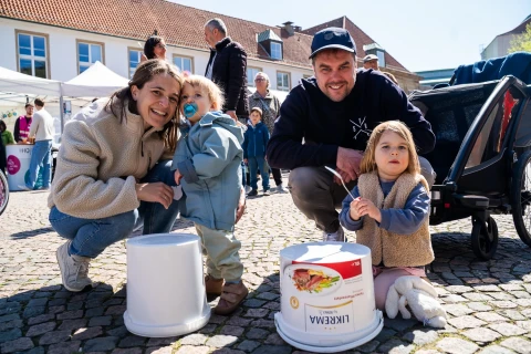 Zwei Erwachsene und zwei Kinder knien auf einem gepflasterten Platz neben zwei wei&szlig;en Plastikeimern. Die Eimer sind als Trommeln genutzt; einer tr&auml;gt ein Etikett mit einer Aufschrift und einem Bild von Lebensmitteln. Im Hintergrund sind Menschen, Marktst&auml;nde und H&auml;user mit roten Ziegeld&auml;chern sichtbar.