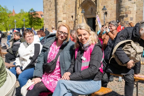 Mehrere Personen sitzen auf Holzsitzb&auml;nken vor einer historischen Steingeb&auml;udefassade mit einem Bogenportal. Zwei Frauen im Vordergrund tragen helle und dunkle Jacken, eine davon ein pinkes Kleid. Menschen im Hintergrund sind teils sitzend, teils stehend. Im Bild verteilt bunte Fahnen, B&auml;ume und Lampenpfosten unter blauem Himmel.