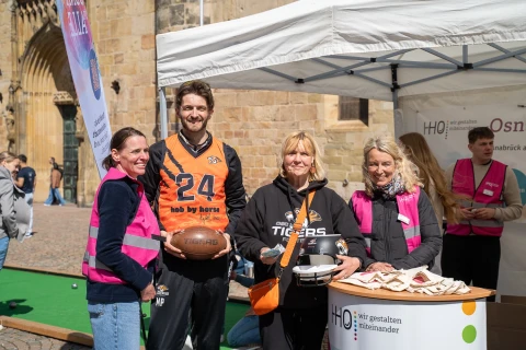 Gruppe von vier Personen unter einem wei&szlig;en Pavillon vor einem historischen Geb&auml;ude. Zwei Personen tragen rosa Warnwesten, eine h&auml;lt einen American Football, eine andere h&auml;lt einen Footballhelm. Ein Tisch im Vordergrund ist mit Stofftaschen und einem Banner mit der Aufschrift &bdquo;HHO wir gestalten miteinander&ldquo; dekoriert.