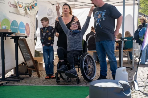 Ein Kind im Rollstuhl streckt beide Arme nach oben. Links steht ein Junge mit verschr&auml;nkten Armen, rechts ein Mann, der nach unten schaut, dahinter eine Frau. Sie stehen unter einem wei&szlig;en Pavillon mit Infotafeln; vorne eine gr&uuml;ne Matte und ein Sitzw&uuml;rfel.