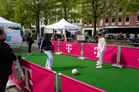 Personen spielen mit einem Ball auf einem rechteckigen Spielfeld mit gr&uuml;nem Kunstrasen, umgeben von pinkfarbenen Absperrungen mit wei&szlig;en "T"-Logos. Im Hintergrund stehen wei&szlig;e Zelte, ein Flipchart und St&auml;nde, begleitet von B&auml;umen und Geb&auml;uden. Ein wei&szlig;er Reisebus und Fahrr&auml;der sind ebenfalls sichtbar.