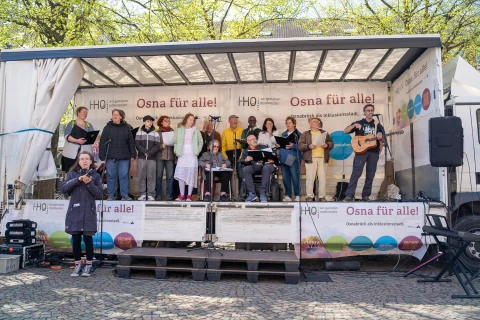 Gruppe von etwa 15 Personen auf einer mobilen B&uuml;hne; eine Person links h&auml;lt Geb&auml;rdensprache, rechts spielt jemand Gitarre. B&uuml;hne dekoriert mit Bannern: "Osna f&uuml;r alle! Osnabr&uuml;ck als Inklusionsstadt". Einige Personen singen, andere halten Textbl&auml;tter, Hintergrund mit B&auml;umen und hellem Himmel.
