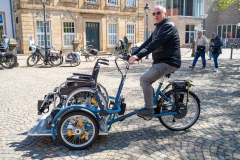 Blaues Dreirad mit einem vorderen Rollstuhlsitz, besetzt von einem Rollstuhl mit schwarzen Polstern und silbernem Gestell. Eine Person in dunkler Kleidung f&auml;hrt das Fahrrad. Kopfsteinpflasterplatz mit Passanten, mehreren Fahrr&auml;dern und historischen Geb&auml;uden im Hintergrund.