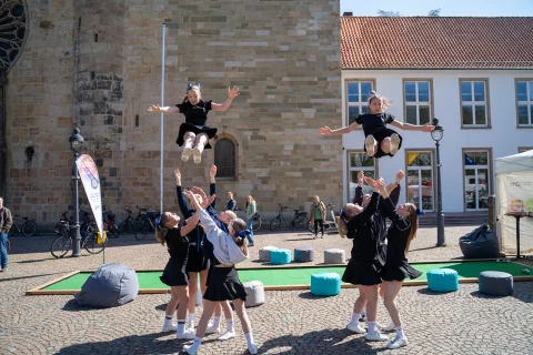Eine Gruppe von Cheerleadern in schwarz-wei&szlig;en Outfits f&uuml;hrt auf einem gepflasterten Platz vor einem Geb&auml;ude akrobatische W&uuml;rfe aus. Zwei Personen befinden sich gleichzeitig in der Luft, umgeben von Teammitgliedern, die sie auffangen. Im Hintergrund ein historisches Geb&auml;ude mit Sandsteinfassade und moderne wei&szlig;e Geb&auml;ude mit roten Ziegeld&auml;chern. Links ein Banner und Fahrr&auml;der, rechts Sitzgelegenheiten auf bunten Sitzs&auml;cken.