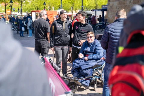 Gruppe von f&uuml;nf Personen im Freien bei sonnigem Wetter, darunter ein Rollstuhlfahrer in blauer Jacke und vier stehende Personen, teilweise in schwarzen Jacken mit Logos. Umgeben von weiteren Passanten, roten und wei&szlig;en Bannern sowie B&auml;umen mit gr&uuml;nem Laub.