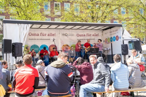 Menschen in bunter Kleidung sitzen und musizieren auf einer erh&ouml;hten B&uuml;hne eines Lastwagens, mit der Aufschrift "Osna f&uuml;r alle! Osnabr&uuml;ck als Inklusionsstadt." im Hintergrund. Vor der B&uuml;hne sitzen Zuschauer auf Holzb&auml;nken. Die Szene ist von gr&uuml;nen B&auml;umen und einem Geb&auml;ude mit Fenstern umrahmt.