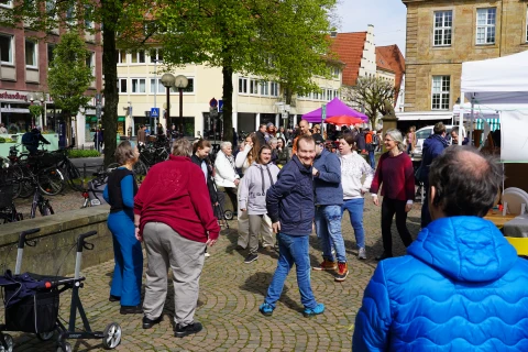 Menschen stehen und gehen auf einem gepflasterten Platz mit B&auml;umen und Fahrr&auml;dern, umgeben von Gesch&auml;ften und Geb&auml;uden mit roten Ziegeld&auml;chern. Rechts sind wei&szlig;e und lilafarbene Marktst&auml;nde mit Sonnenschirmen sichtbar. Im Vordergrund ein Rollator und eine Person in einem roten Pullover.