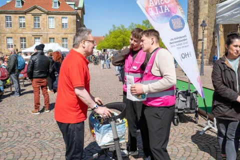 Mann in rotem T-Shirt steht vor zwei Personen in pinken Westen, die eine Unterhaltung f&uuml;hren. Der Mann lehnt an einem beladenen Rollator. Rechts neben den pinken Westen ist eine wei&szlig;e Fahne mit Aufschrift sowie ein QR-Code zu sehen. Pflastersteine bedecken den Boden, im Hintergrund stehen Personen, Marktst&auml;nde und historische Geb&auml;ude mit roten Ziegeld&auml;chern.