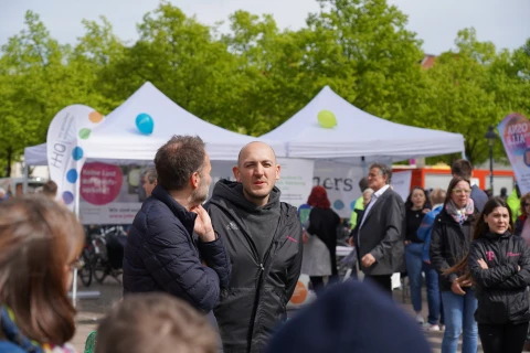 Personen unterhalten sich auf einem &ouml;ffentlichen Platz vor wei&szlig;en Pavillons mit Informationsst&auml;nden, bedruckten Bannern und Luftballons in Gelb, Blau und Gr&uuml;n. Im Hintergrund gr&uuml;ne B&auml;ume und kleinere Gruppen weiterer Personen. Ein gro&szlig;es Banner zeigt den Schriftzug &bdquo;Keine Last mehr auf dem Wertstoff&ldquo;.