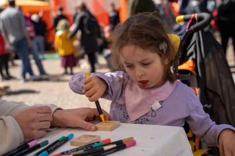 Kind in lila Oberteil sitzt an einem Tisch und h&auml;lt gelben Stift, greift Holzst&uuml;ck mit ausgestanztem Muster. Tisch mit wei&szlig;en Tuch, mehrere Stifte in verschiedenen Farben. Hintergrund: unscharfe Personen in bunter Kleidung, orangefarbene und gr&uuml;ne Zelte.