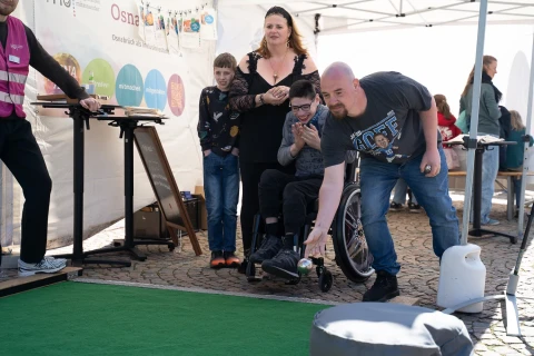 Eine Mutter mit zwei Kindern und einem Mann neben einem Kind im Rollstuhl steht vor einem Mini-Golfplatz mit gr&uuml;nem Teppich. Der Mann h&auml;lt einen Golfschl&auml;ger und hilft dem Kind, einen Ball zu schlagen. Im Hintergrund ein wei&szlig;es Zelt mit Informationsplakaten und einige Personen.