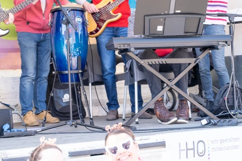 Menschengruppe auf einer B&uuml;hne. Vier Personen spielen Musikinstrumente: E-Gitarre, Bass, Congas und Querfl&ouml;te. Ein Keyboard steht rechts. Hinter ihnen ein Banner mit &bdquo;Osna f&uuml;r alle!&ldquo; und Texten zur Inklusion. Unten vor der B&uuml;hne sitzen andere Personen, unscharf durch helle Belichtung, auf einem Schild steht &bdquo;HHO &ndash; wir gestalten miteinander&ldquo;.