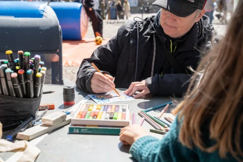 Person mit schwarzer Jacke und Kappe zeichnet mit einem orangefarbenen Bleistift auf einem kleinen wei&szlig;en Papier an einem Tisch im Freien. Darauf diverse Malutensilien wie bunte Pastellkreiden, Stifte und Marker. Im Vordergrund der R&uuml;cken einer weiteren Person, im Hintergrund unscharfe Details einer Veranstaltung im Freien.