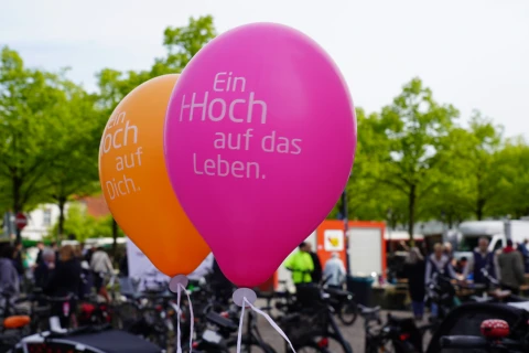 Zwei heliumgef&uuml;llte Luftballons mit aufgedrucktem Text; der linke orangefarbene Ballon tr&auml;gt die Aufschrift "Ein Hoch auf Dich.", der rechte pinkfarbene Ballon die Aufschrift "Ein Hoch auf das Leben.". Im Hintergrund unscharfe Darstellung eines belebten Platzes mit Fahrr&auml;dern, Personen und gr&uuml;n belaubten B&auml;umen.
