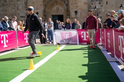 Ein Mann mit grauem Baseballcap und schwarzer Jacke f&uuml;hrt auf einem schmalen gr&uuml;nen Kunstrasenpfad mit wei&szlig;en Linien einen Fu&szlig;ball an gelben H&uuml;tchen vorbei. Mehrere Menschen schauen zu, einige stehen hinter Werbebannern mit pinkem Hintergrund und wei&szlig;em "T"-Logo. Im Hintergrund eine sandfarbene Geb&auml;udefassade mit Rundbogenportal.