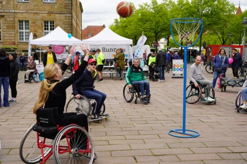 Frau im Rollstuhl mit schwarzem Shirt und blonden Haaren wirft Basketball in einen blauen Basketballkorb, umgeben von weiteren Personen in Rollst&uuml;hlen. Im Hintergrund wei&szlig;e Zelte, eine steinerne Geb&auml;udefassade, gr&uuml;ne B&auml;ume und mehrere Menschen. Holpriger Platz mit grauen Pflastersteinen.