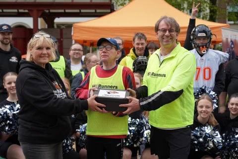 Eine Gruppe von Personen in Sportkleidung steht vor einem orangen Pavillon, darunter drei Personen im Vordergrund in gelber Runners-Sportkleidung, die gemeinsam ein schwarzes rechteckiges Objekt halten. Hintergrund mit weiteren Menschen und Cheerleaderinnen mit Pompons.